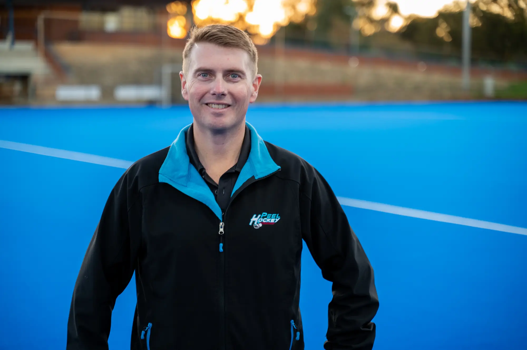 Person stands on a bright blue hockey field wearing a black jacket with blue trim. White field markings cross the turf, with sports facilities and trees softly blurred in the background.